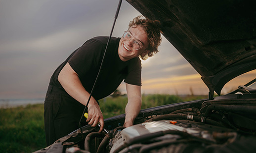 Quinn Legg stands near a car with an open hood and looks to his right while holding a wrench and smiling.