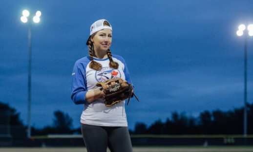    A woman wearing a Toronto Blue Jays shirt, a baseball cap and baseball glove stands in a baseball field under bright stadium lights.