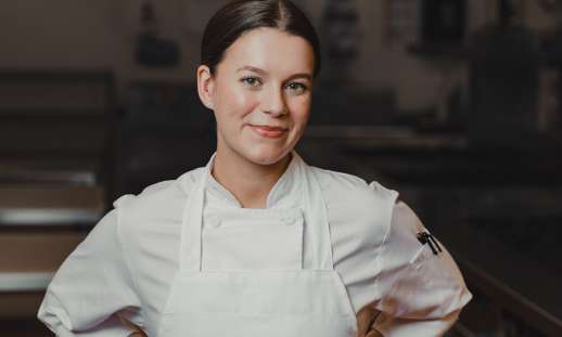 Olivia Sewell standing in a culinary kitchen in cooking attire, smiling at the camera.