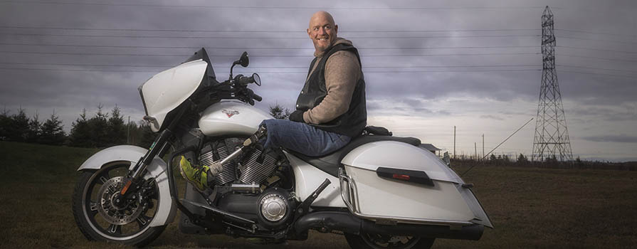 A man smiles while sitting astride a white motorcycle. He is wearing jeans, a brown sweater and a vest. His leg is a prosthetic, and he is wearing a bright green sneaker. There is a large power line in the background.