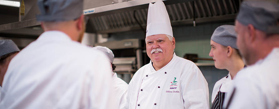 An man wearing a chef's jacket and tall chef's hat stands in a commercial kitchen. Five chefs in pill-box caps and white chef’s jackets are gathered around the other man and appear to be listening to something he is saying. Jars can be seen on the workspace in front of them.
