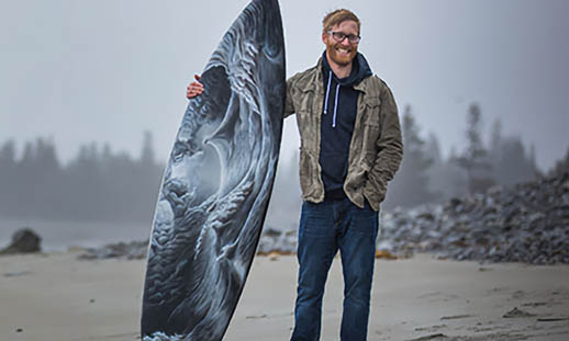 A man stands on a beach holding a surfboard painted with waves. He is wearing a jacket, sweater and jeans. There is mist and trees in the background.