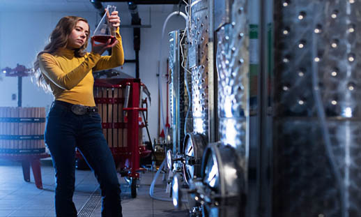 A woman in a yellow sweater looks closely at a glass vessel holding red wine. She is in a wine-making area.