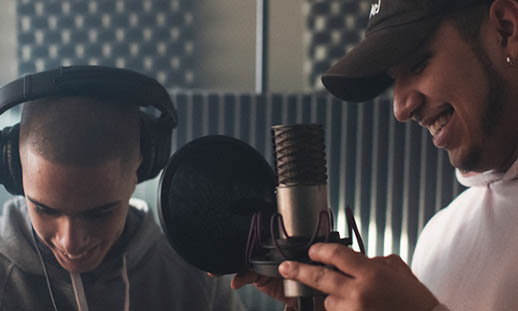Two men smile while standing close to a professional-looking microphone. They are in a sound booth.