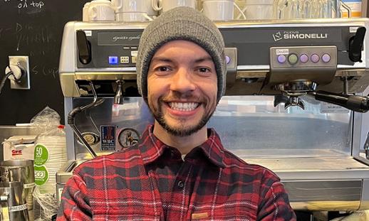 Leo stands in front of an industrial coffee/espresso maker, wearing a beanie and plaid flannel shirt.