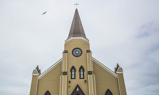 The top half of St. Mary's Polish Parish is seen against a cloudy sky. On the center steeple a metal cross can be seen. A bird is flying on the left.
