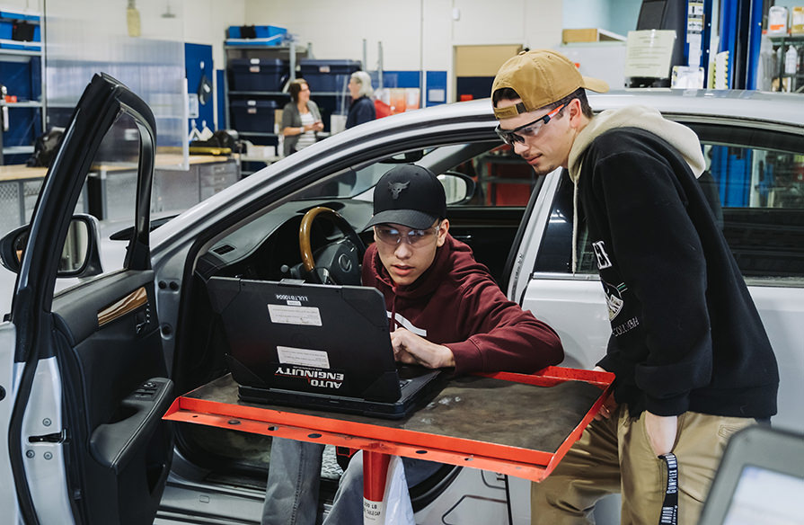 Two students look at a computer while working on a car in an auto shop setting.