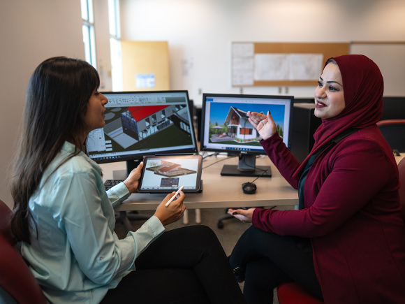 Two female students look at renderings on their computer screens.