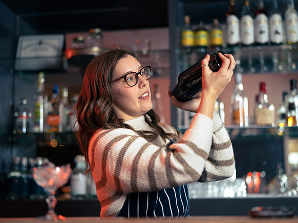 A woman in a striped sweater and glasses shakes a cocktail shaker while standing behind a restaurant bar.