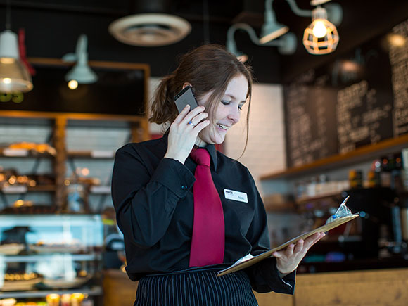 A student in a uniform talks on a cell phone while looking at a clipboard in a restaurant setting.