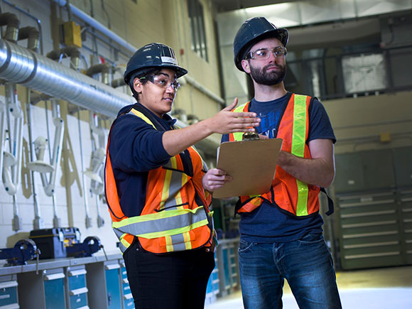Two people in hard hats, safety glasses and safety vests look at a clipboard in a workshop.
