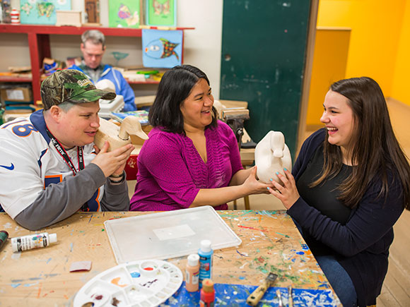 A disability support worker smiles as two people show her an artistic project they are working on.
