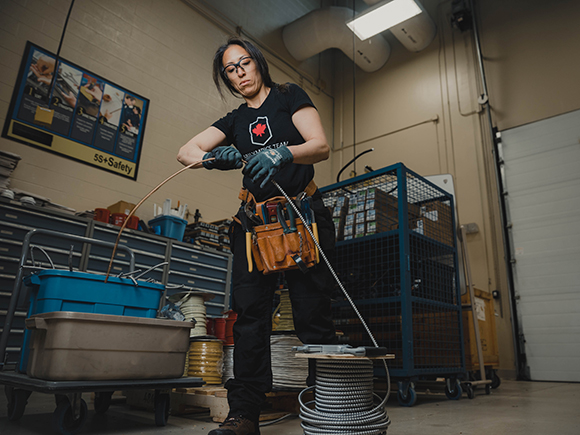 A woman standing in front of a work bench with many tools wearing safety glasses and gloves adjusts a thick, metal wire.