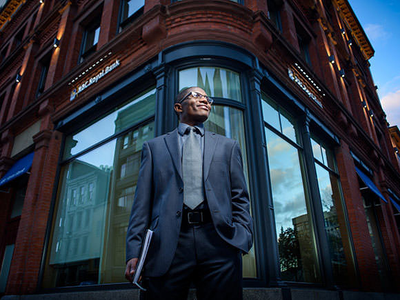 A man with glasses, wearing a suit stands outside a city building.