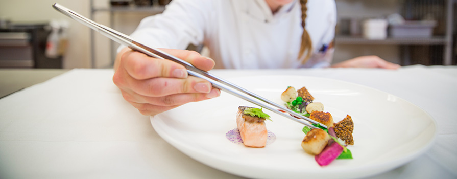 A close-up photo of a chef’s hands carefully placing a variety of bite-sized foods on a white plate using long silver tweezers.