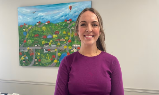 Brynn Budden smiles and wears a purple shirt. She stands in front of a colourful and vibrant painting.