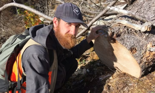 A white man with a think red beard stoops down next to a cut tree trunk on the ground.