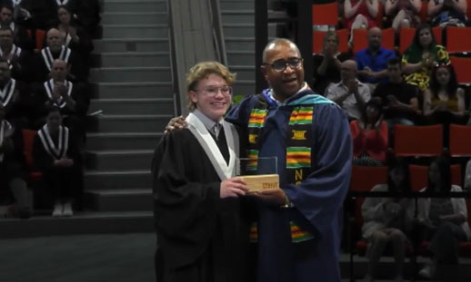 Two individuals stand on the stage, one receiving their graduation certificate and the other handing it to them.