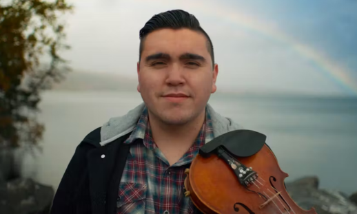 Morgan Toney of We'koqma'q First Nation standing outside. He's holding a fiddle and there's a rainbow peaking through in the fog behind him.