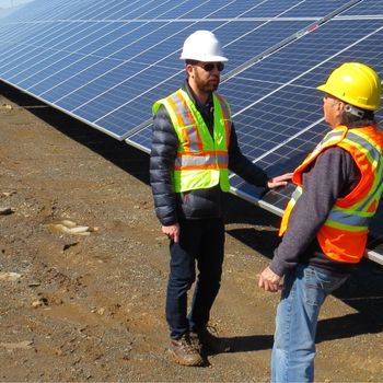 Two men wearing hard hats and safety vests, while they're talking in front of solar panels on the ground. 