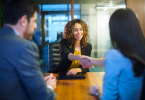 Two women and a man seated at a boardroom table and the women are shaking hands.