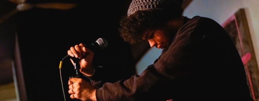 A closeup of a Black man holding a mic and its stand on stage before performing spoken word.