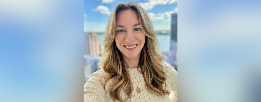 Photo of Niecole Killawee in front of window with background showing a bright cityscape with tall buildings and a body of water under a blue sky with scattered clouds.