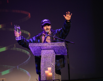 Musician P’tit Belliveau on stage standing at a podium to give their award acceptance speech.