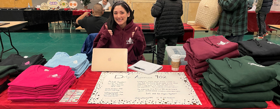 Tyra Paul's booth at a craft fair inside a school's gym. She sitting at a table filled with t-shirts and sweaters from her business, Drip Avenue 902.