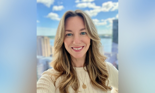 Photo of Niecole Killawee in front of window with background showing a bright cityscape with tall buildings and a body of water under a blue sky with scattered clouds.