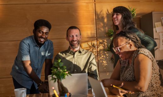 Two men and a woman sit at a boardroom table, talking to each other while using laptops and notebooks. Another woman is standing next to the table, talking with the group.