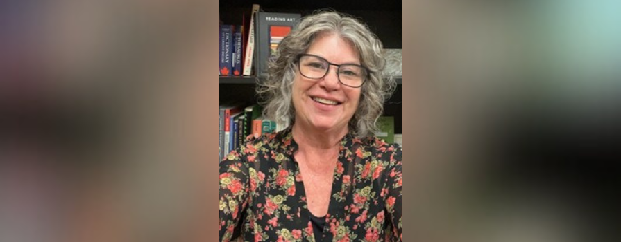 Pam Eakin sitting in front of a bookshelf filled with books, wearing a black floral-patterned top.