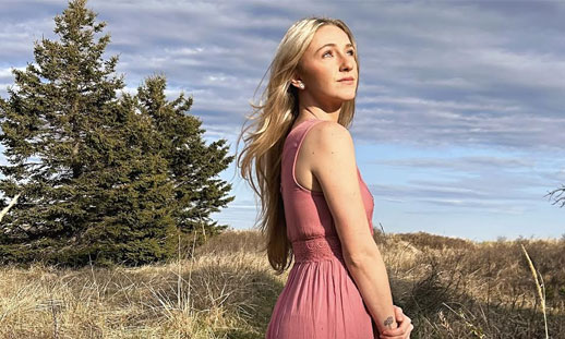 A woman with blonde hair and a pink dress is staring up to the sky while standing in a field.