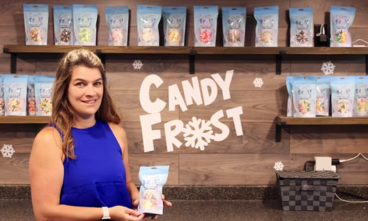 Candice Mugford, owner of Candy Frost, smiling and holding a bag of her candy in front of a wall with shelves filled with her products.