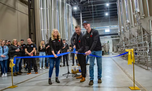 Pictured is co-founders Olivia Giffen, left, Riley Giffen, and Liam Giffen cutting a blue ribbon in their new Coldstream Clear brewery. A group of people are watching and smiling.