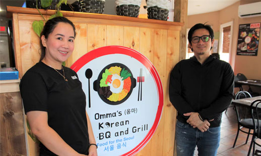 Two restaurant owners, male and female stand in the entrance of their new location in front of their logo that reads "Omma's Korean BBQ and Grill."