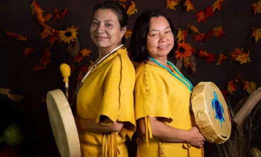 We’koqma’q sisters Ella and Eva Nicholas standing back-to-back in traditional wear while each holding a drum.