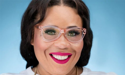 A woman wearing pink lipstick, light pink framed glasses and a white top is photographed, smiling with a blue background.