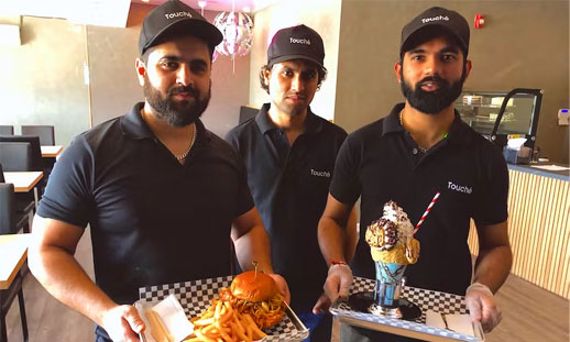 Three men hold items from their new restaurant, including a milkshake with tons of toppings and a butter chicken sandwich with fries.