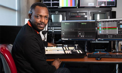 A man in a headset is seated at a recording studio desk. There are monitors and microphones seen.