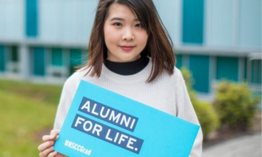 Asian woman standing outside a campus and holding a blue sign that says "Alumni for Life"