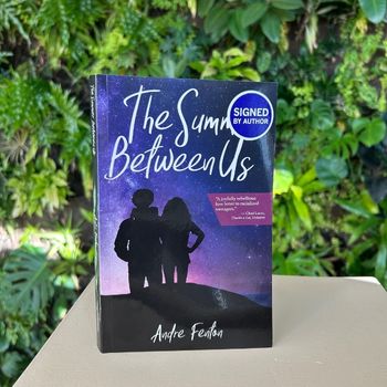 A closeup of the book, "The Summer Between Us" on a table with a plant wall in the background.