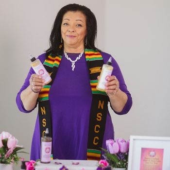 An African Canadian woman smiles as she holds of her natural products, standing behind a display of decorative jars on a table.