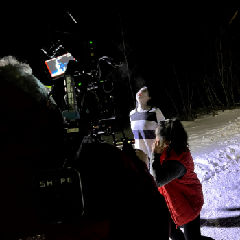 A videographer recording a scene in the snow with a ghost-like woman screaming. A female set working is standing near the actress.