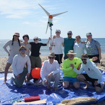 A group photo of 11 people, both women and men, standing and kneeling on a blue tarp on a beach in France with their floating structures for wind turbines.