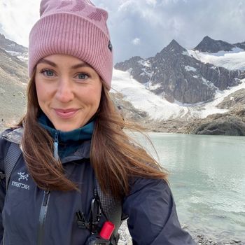 A selfie of a woman wearing a pink toque and light jacket while hiking in front of mountains.