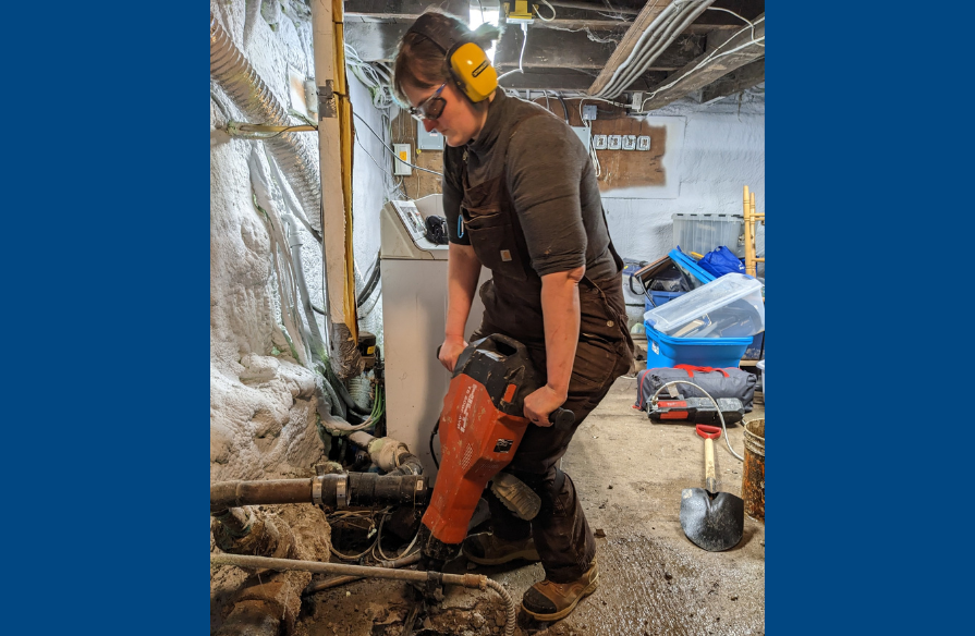 A woman wearing PPE and using a jackhammer to install a sump pump in a basement.