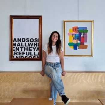 A brunette woman sitting crossed leg on the back of a couch in front of two visual art pieces. She's smiling at the camera.