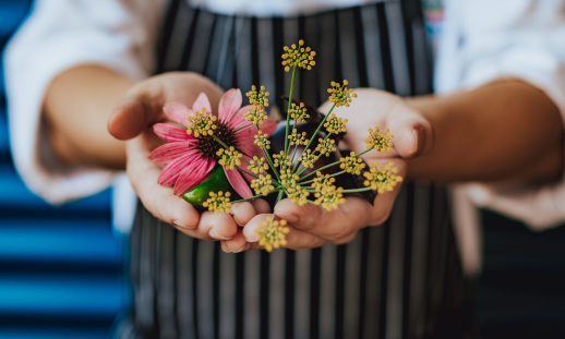 Close-up photo of open hands holding flowers with the background showing the individual wearing a cooking apron.