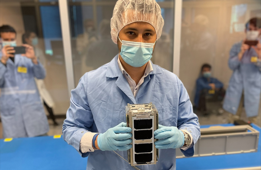 Arad in a lab holding the LORIS satellite with lab workers in the background behind a glass wall.